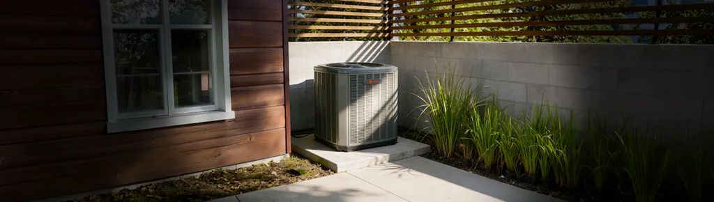 Um condensador Trane sobre uma base de concreto ao lado de uma casa revestida de cedro, com gramíneas ornamentais ao redor